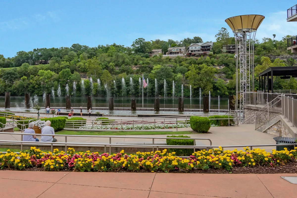 This image features lively fountains, vibrant flower beds, a river, and people strolling along a scenic area. The scene captures the essence of a dynamic downtown, with visible pedestrian activity and a backdrop of greenery and water that hints at waterfront attractions.