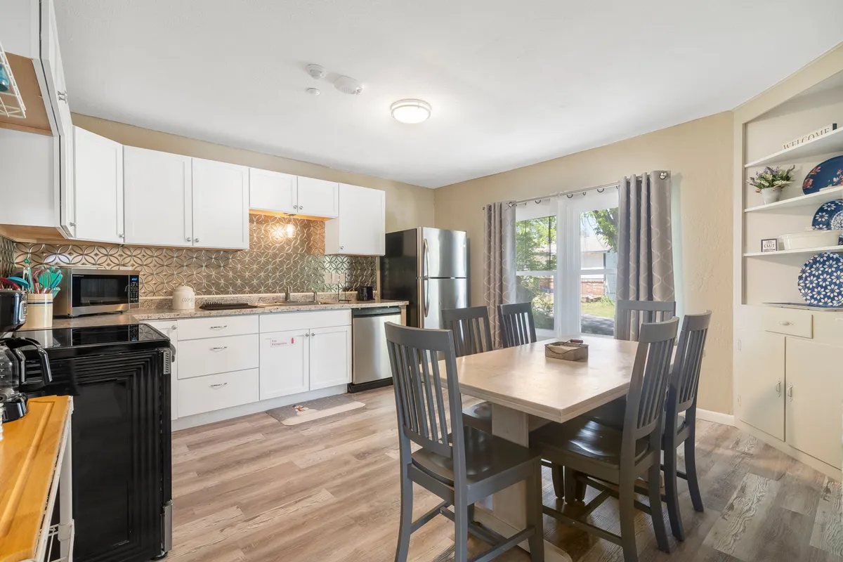 A bright dining area with a spacious dining table, kitchen island, large windows allowing in abundant sunlight, and decorative shelves. The open layout and warm tones invite guests to gather for meals or lively conversation, reinforcing a sense of community.