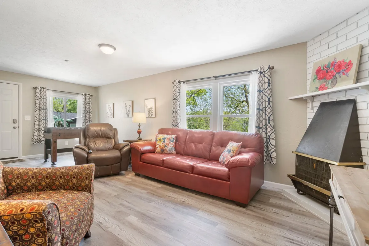 A cozy living room featuring a red leather sofa, a colorful accent chair, large windows streaming in natural light, decorative wall art, and a standing lamp. The space is thoughtfully arranged to encourage both relaxation and conversation, with soft textures and inviting décor elements.