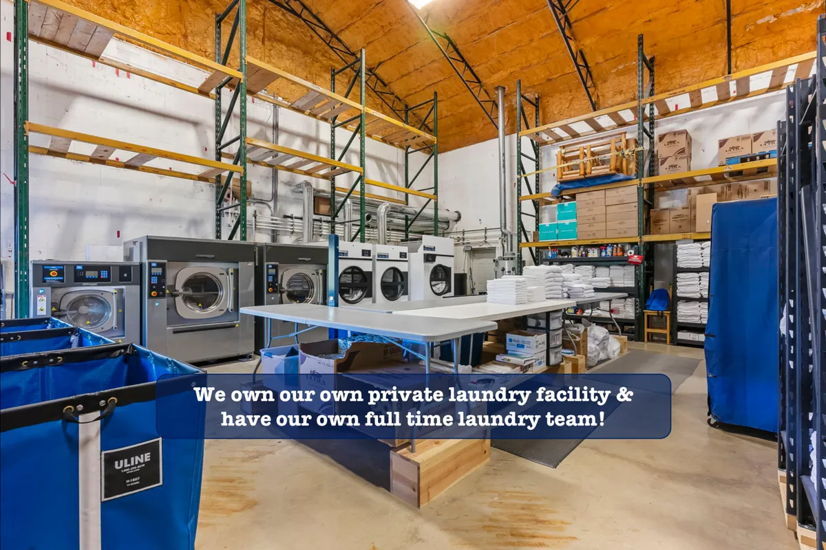 A bright, organized laundry room featuring industrial laundry machines, a folding table, storage shelves, and laundry bins. The space is clearly built for efficiency, showcasing neatly arranged detergents and folded linens, reflecting its professional-grade cleaning capabilities.