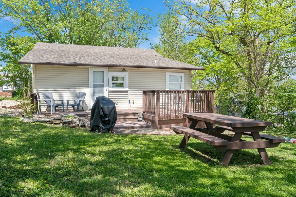 A welcoming backyard scene features a small house with a wooden deck, a picnic table, a grill, and plenty of greenery. The relaxed setting is perfect for BBQs, outdoor dining, and family gatherings, inviting guests to enjoy the outdoors in comfort.