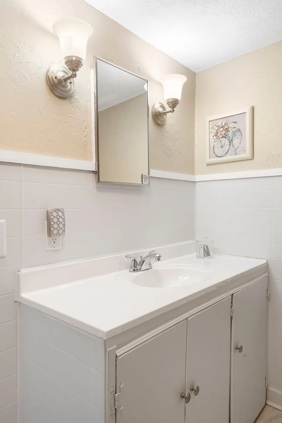 A serene bathroom interior featuring a sink beneath a well-lit mirror, elegant light fixtures, and tasteful wall art. Morning light pours in, adding a sense of calm and freshness—inviting moments of self-care and relaxation.
