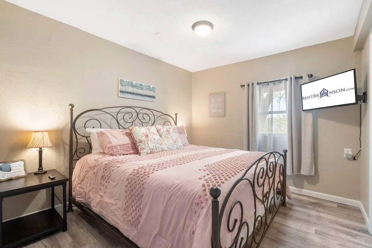 A well-lit bedroom featuring a bed with decorative pillows, a nightstand with a lamp, a television mounted on the wall, and a window dressed with curtains. Soft natural light streams in, creating a cozy and tranquil atmosphere ideal for rest and relaxation.